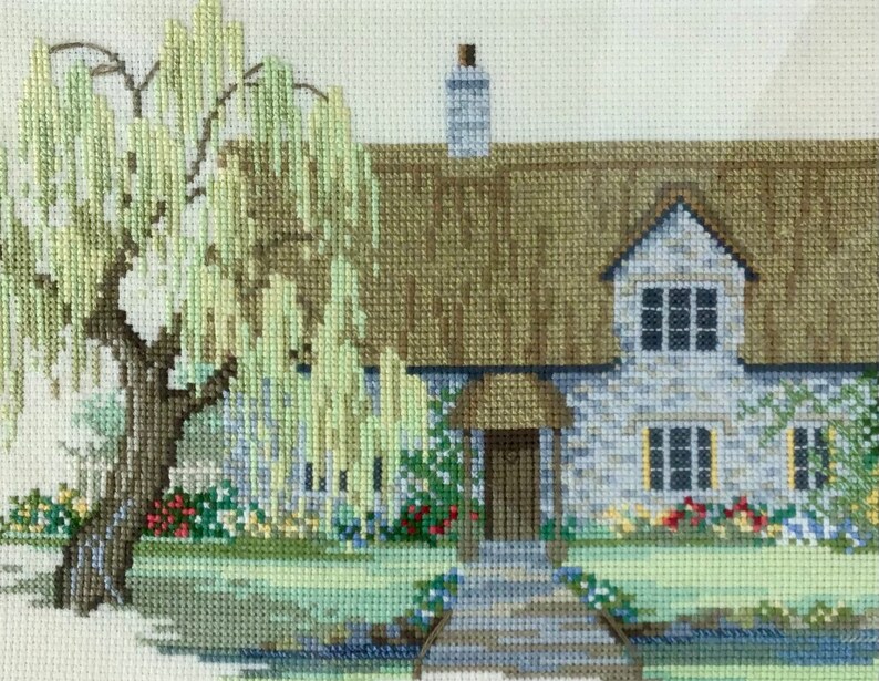 K&ouml;nnte beinhalten: Eine Kreuzstich-Stickerei, die ein Cottage mit einem Strohdach, einer Steinfassade und einer Trauerweide im Vordergrund zeigt. Das Cottage hat eine braune T&uuml;r und Fenster mit wei&szlig;en Rahmen. Die Trauerweide hat gr&uuml;ne Bl&auml;tter und &Auml;ste, die bis zum Boden h&auml;ngen.