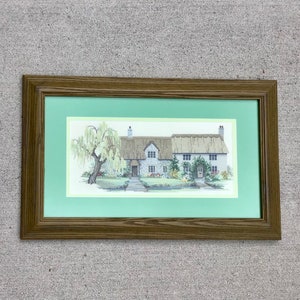 K&ouml;nnte beinhalten: Ein gerahmtes Kreuzstich-Bild, das ein wei&szlig;es Cottage mit Strohdach zeigt, umgeben von Gr&uuml;n und einem Baum. Das Cottage hat eine Haust&uuml;r und Fenster, und das Bild ist in einem braunen Holzrahmen mit gr&uuml;nem Passepartout gerahmt.