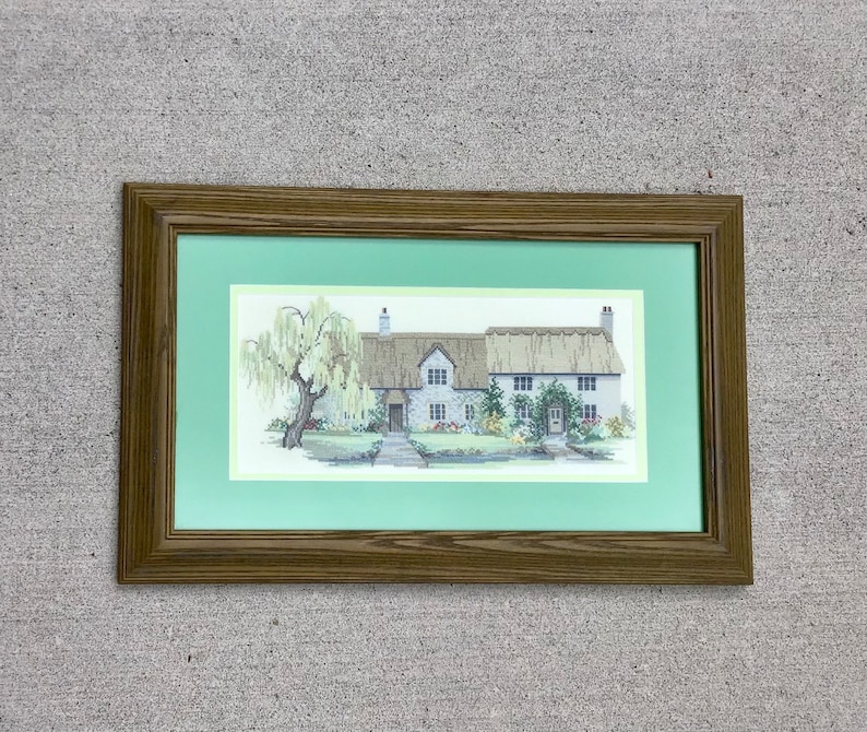 K&ouml;nnte beinhalten: Ein gerahmtes Kreuzstich-Bild, das ein wei&szlig;es Cottage mit Strohdach zeigt, umgeben von Gr&uuml;n und einem Baum. Das Cottage hat eine Haust&uuml;r und Fenster, und das Bild ist in einem braunen Holzrahmen mit gr&uuml;nem Passepartout gerahmt.