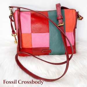 May include: A patchwork Fossil crossbody bag in shades of red, pink, orange, and teal. The bag has a long, adjustable strap and a gold-tone key charm. The text "Fossil Crossbody" is at the bottom. The bag is made of leather.