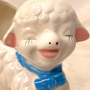 May include: A vintage ceramic lamb planter with a smiling face, closed eyes, and a blue bow. The lamb has pink ears and a red nose. The planter is white with a glossy finish.