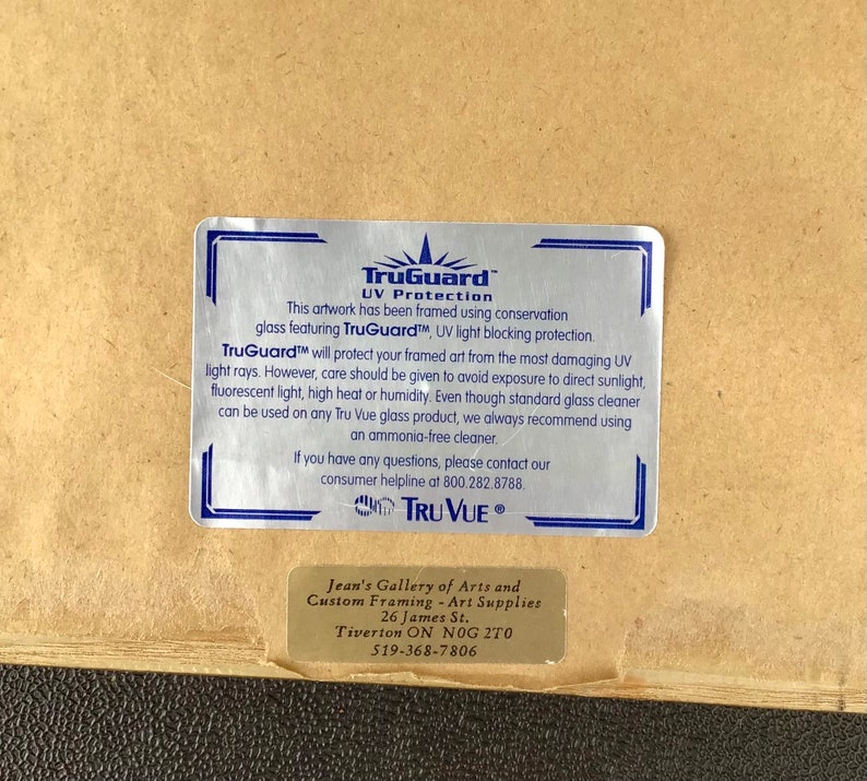 K&ouml;nnte beinhalten: Ein TruGuard UV Protection-Etikett auf braunem Hintergrund. Das Etikett ist silberfarben mit schwarzem Text und einem TruVue-Logo. Der Text lautet: "Dieses Kunstwerk wurde mit einem Konservierungsglas mit TruGuard-UV-Lichtblockierung gerahmt. TruGuard sch&uuml;tzt Ihre gerahmte Kunst vor den sch&auml;dlichsten UV-Strahlen. Es sollte jedoch darauf geachtet werden, dass keine direkte Sonneneinstrahlung, fluoreszierendes Licht, hohe Hitze oder Feuchtigkeit auftreten. Obwohl Standard-Glasreiniger f&uuml;r alle Tru Vue-Glasprodukte verwendet werden k&ouml;nnen, empfehlen wir immer die Verwendung eines ammoniakfreien Reinigers. Wenn Sie Fragen haben, wenden Sie sich bitte an unsere Kundenhotline unter 800.282.8788. TruVue."
