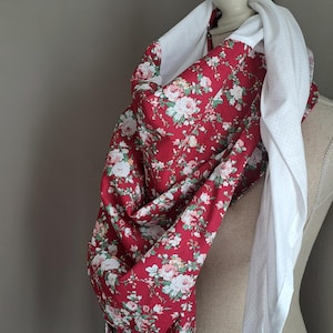 Puede incluir: Una bufanda roja floral con rosas blancas y rosas y hojas verdes está drapeada sobre un maniquí. Una bufanda blanca con pequeños lunares grises está drapeada sobre el cuello del maniquí. La bufanda está hecha de tela suave.