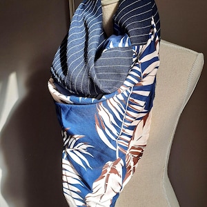 Può includere: Una sciarpa ad anello a motivi multipli caratterizzata da un tessuto blu scuro con sottili righe bianche e una sezione a contrasto con una stampa di foglie tropicali nei toni del blu, bianco e marrone, drappeggiata su un manichino.