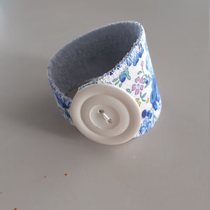 Könnte beinhalten: Ein weißes Stoffarmband mit einem blauen Blumenmuster und einem weißen Knopfverschluss.