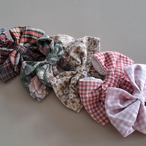 Könnte beinhalten: Eine Sammlung von Stoff-Haarschleifen in verschiedenen Mustern und Farben. Die Schleifen umfassen Blumen-, Karo- und Vichy-Designs in Rosa-, Rot-, Grün- und Brauntönen. Accessoires für alle Altersgruppen.