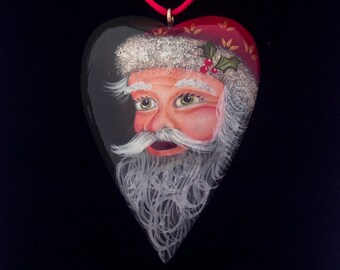 May include: A black heart-shaped ornament with a painted image of Santa Claus. Santa has a white beard and mustache, a red hat with green holly, and a white fur trim. The ornament has a red cord for hanging.