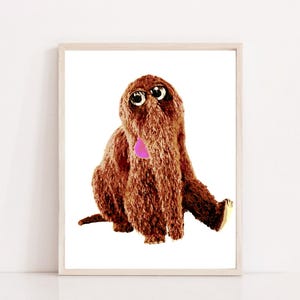 May include: A brown furry puppet with large eyes and a pink tongue sticking out. The puppet is sitting on a white background.