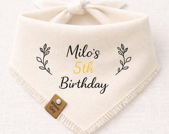 May include: Cream-colored dog bandana with the text "Milo's 5th Birthday" in black and yellow. The bandana has a knotted tie and is decorated with leaf sprigs. A small brown tag with the text "Whitney Grey" is attached.