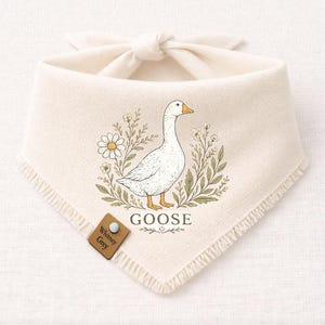 May include: Cream-colored pet bandana with a white goose illustration surrounded by floral elements. The word "GOOSE" is printed below the goose. A small brown tag with the text "Whitney Grey" is attached to the bandana.