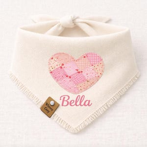 May include: Cream-colored pet bandana with a patchwork heart design in shades of pink. The name "Bella" is printed in pink script below the heart. A small brown tag with the text "Whitney Grey" is attached.