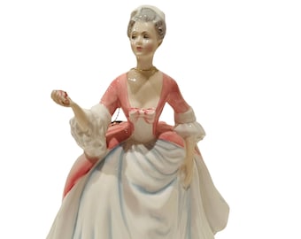 以下が含まれることがあります： 白い帽子をかぶったピンクと白のドレスを着た女性の磁器製フィギュア。彼女は手に小さな赤い物を持っています。