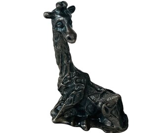 May include: A small, silver-colored giraffe figurine with a textured surface. The giraffe is sitting with its head turned to the side.