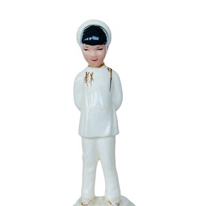 May include: A white porcelain figurine of a person wearing a white hat and traditional clothing. The figurine has black hair and a gold design on the clothing.