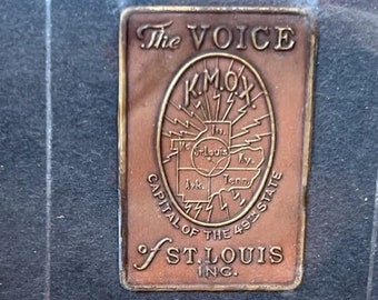 Può includere: Una placca in bronzo vintage con il testo "The Voice KMOX Capital of the 49th State of St. Louis Inc." e una mappa degli Stati Uniti con un design a forma di fulmine.