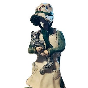 May include: A ceramic figurine of a woman wearing a green dress and a floral hat, holding a small gray cat. The figurine is standing on a green base with floral details.