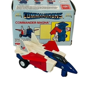May include: A red, white, and blue toy jet fighter plane with the words "COMMANDER MAGNA" on the box. The toy is in the shape of a jet fighter and has a red, white, and blue color scheme. The toy is made of plastic and has a simple design.