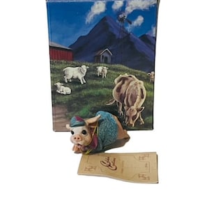 May include: A ceramic figurine of a pig wearing a blue and green coat and hat, with a brown base. The figurine is in front of a box with a rural landscape scene featuring sheep, cows, and a barn.