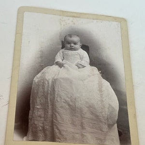 May include: Vintage black and white photograph of a baby seated in a chair, wearing a long white dress. The photograph is mounted on a card with the text "Hatch & Waite, Burlington, Wis."