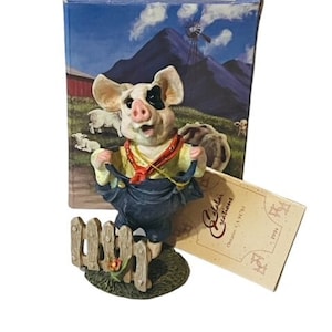 May include: A white and black pig figurine wearing blue overalls and a red bandana, standing by a wooden fence. The figurine is part of a collection called "Calabar Creations".