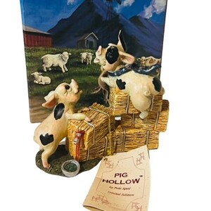 May include: A figurine of two pigs, one standing on hay bales and the other looking up at it. The figurine is made of painted resin and has a rustic, country feel. The figurine is packaged in a box with a blue background and a picture of a farm scene. The box also has the text "Pig Hollow" on it.