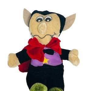 Könnte beinhalten: Ein Plüschtier-Vampir mit einem roten Umhang, einer lila Weste und einem Stern auf der Weste. Das Spielzeug hat ein braunes Gesicht mit einer großen Nase und Zähnen.