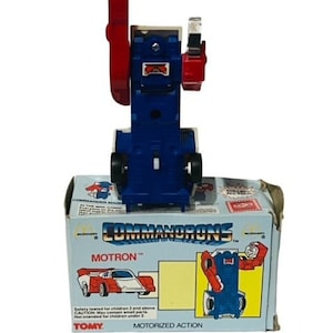 May include: A blue and red toy robot in car form, called "MOTRON", from the "COMMANDRONS" toy line. The robot is in its car form, with wheels visible. The box is white with blue and red accents. The box has the text "COMMANDRONS" and "MOTRON" on it. The box also has the text "MOTORIZED ACTION" on it.