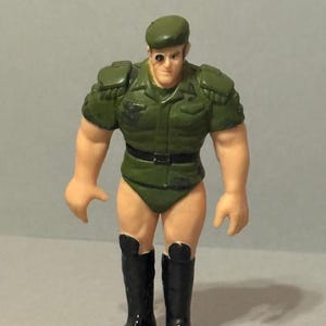 Könnte beinhalten: Vintage Actionfigur eines Soldaten in grüner Uniform, schwarzen Stiefeln und einem grünen Barett. Die Figur hat einen muskulösen Körperbau und eine Augenklappe. Das Spielzeug steht aufrecht.