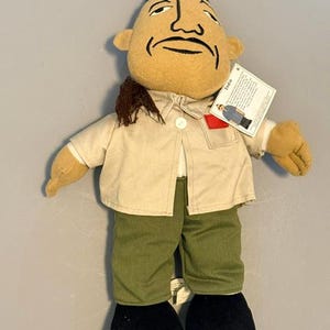 May include: A plush doll with a tan face, black hair, and a mustache, wearing a beige button-down shirt with a red patch on the chest pocket and olive green pants. The doll has black shoes and a white tag attached to the shirt collar labeled Indio.