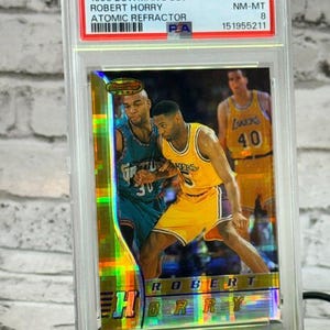 Op de afbeelding: Een 1996 Bowman's Best Robert Horry Atomic Refractor basketbalkaart, beoordeeld als NM-MT 8 door PSA. De kaart toont Robert Horry in een geel Lakers-uniform en een andere speler in een blauwgroen Grizzlies-uniform, tegen een holografische achtergrond.