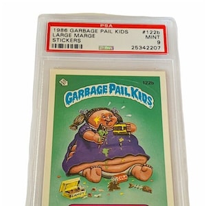 Puede incluir: Una tarjeta de intercambio de Garbage Pail Kids calificada que presenta a Large Marge, un personaje con un vestido morado y una cara sucia, rodeado de dulces y otros art&iacute;culos. La tarjeta est&aacute; calificada con un 9 por PSA y tiene el n&uacute;mero #122b.