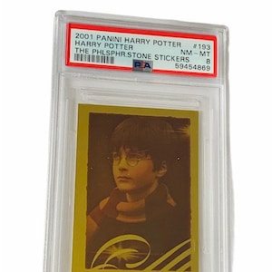 May include: A graded trading card featuring Harry Potter from the 2001 Panini Harry Potter: The Philosopher's Stone Stickers set. The card is in near mint condition and has a gold background with a black and white striped border. The card features a close-up image of Harry Potter with his glasses on and a scarf around his neck.