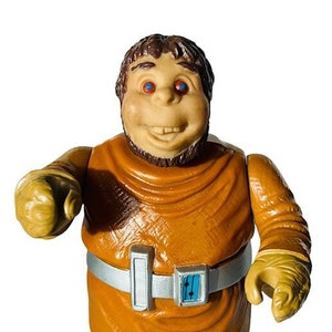 May include: A brown and orange plastic action figure of a creature with a large head, brown hair, and red eyes. The figure is wearing a brown robe with a silver belt and a blue buckle. The figure is holding its hands out in front of it.
