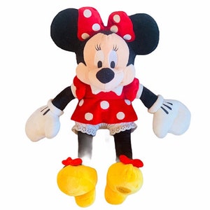 May include: A plush toy of Minnie Mouse wearing a red dress with white polka dots and a red bow with white polka dots. She has black ears and a black nose. Her shoes are yellow with red bows.