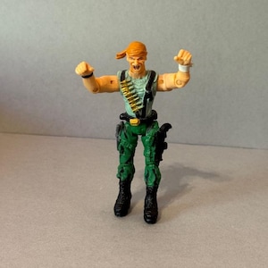 May include: A small action figure of a soldier in a combat pose. The figure is wearing a green and tan uniform with a bandolier of bullets, black boots, and a red bandana. The figure has its arms raised in a celebratory gesture.