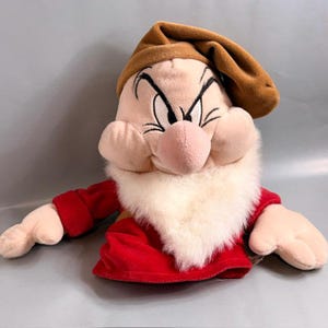 May include: A plush toy of the character Grumpy from Snow White, featuring a tan hat, a large pink nose, thick black eyebrows, and a long white faux fur beard. The character wears a red long-sleeved shirt and has light skin-toned plush hands.