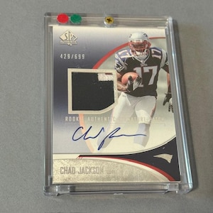 May include: A Chad Jackson rookie card in a protective case. The card features a player in a Patriots uniform, a jersey swatch, and an autograph. The card is numbered 429/699.