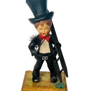 Puede incluir: Figura de un deshollinador, con sombrero de copa negro, chaqueta y pantalón, con pajarita roja. La figura sostiene una escalera y está sobre una base de madera con una escena pintada.