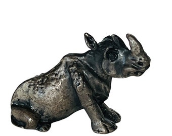 May include: A small, silver-colored rhinoceros figurine. The rhinoceros is sitting on its haunches with its head turned to the side. The figurine is detailed with textured skin and a horn.