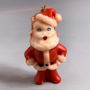 Puede incluir: Una vela de cera moldeada con forma de Santa Claus, que presenta un traje rojo con ribetes blancos, un gorro rojo a juego, una barba blanca y ojos azules. La figura está de pie con las manos en la cintura, una mecha en la parte superior y boca roja.