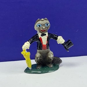 May include: A small, grey figurine of an animal wearing a black tuxedo, a red bow tie, and a top hat. The figurine is holding a yellow umbrella and is standing on a green base.