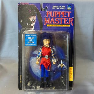 Puede incluir: Una figura de acción de plástico de Jester de la serie de películas Puppet Master. La figura lleva un traje de bufón rojo y azul con un patrón a cuadros. La figura sostiene un arma en su mano derecha. La figura está empaquetada en un blíster de plástico con un fondo azul y amarillo. El empaque incluye el texto "Puppet Master" y "The Action Figure Series".