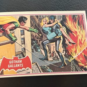 Könnte beinhalten: Eine Vintage-Comic-Sammelkarte mit Batman und Robin. Die Karte hat ein rotes und schwarzes Fledermauslogo mit dem Text "Gotham Gallants". Batman ist in einem blauen Anzug zu sehen, trägt eine Frau in einem blauen Kleid, während Robin in einem roten und grünen Kostüm an einem Seil schwingt.