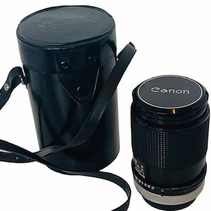 May include: Black Canon camera lens and a matching black leather case. The lens has a silver ring and a black cap with the Canon logo. The case is cylindrical with a shoulder strap. The lens is a 50mm f/1.8.