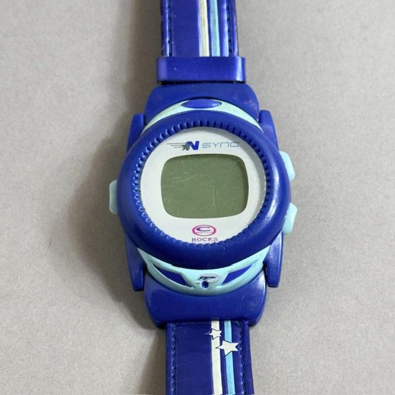 Puede incluir: Reloj digital azul con una correa azul a juego. La esfera del reloj tiene el texto "N SYNC" y "ROCKS". La correa presenta rayas blancas y azul claro y detalles de estrellas. El reloj es de color azul vibrante.