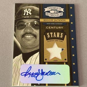 Puede incluir: Una tarjeta de béisbol con Reggie Jackson de los Yankees de Nueva York. La tarjeta incluye una foto de Jackson, el texto "THROWBACK 20 THREADS 04" y un parche en forma de estrella de sus pantalones usados en el juego. La tarjeta está firmada.