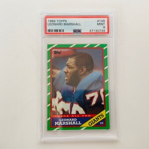 Op de afbeelding: Een 1986 Topps voetbalkaart met Leonard Marshall, een defensive end voor de New York Giants. De kaart is in mint conditie en heeft een groene, witte en rode rand. De kaart toont een foto van Marshall in zijn uniform, met de tekst "TOPPS ALL PRO" en "GIANTS" gedrukt op de kaart.