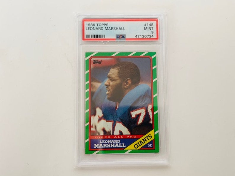 Op de afbeelding: Een 1986 Topps voetbalkaart met Leonard Marshall, een defensive end voor de New York Giants. De kaart is in mint conditie en heeft een groene, witte en rode rand. De kaart toont een foto van Marshall in zijn uniform, met de tekst "TOPPS ALL PRO" en "GIANTS" gedrukt op de kaart.