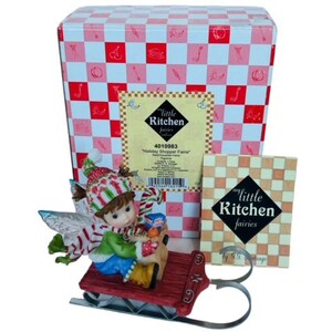 May include: A figurine of a little kitchen fairy wearing a red and white striped hat and a green scarf, riding a red sled with a silver base. The fairy is holding a basket of fruit and a candy cane. The figurine is packaged in a box with a red and white checkered pattern and the words "My Little Kitchen Fairies by S.T. Santiago".