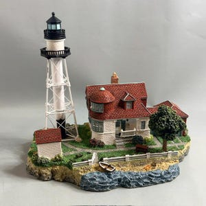 May include: A miniature lighthouse figurine featuring a tall white tower with a black lantern room, positioned next to a white house with a red shingled roof. The scene includes a small boat, a green tree, and a white fence on a rocky base.